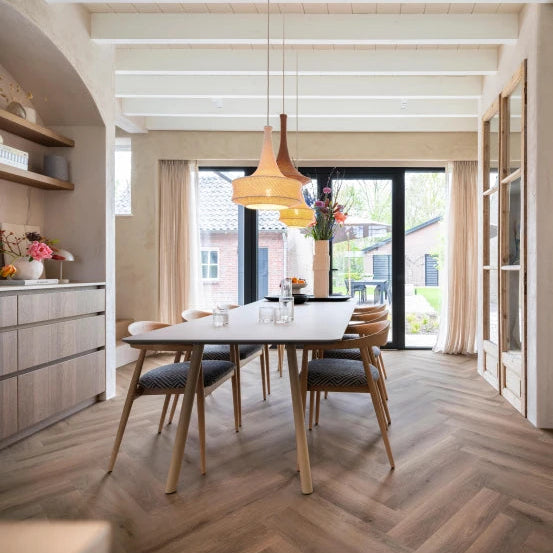 lichtgrijze vloer met visgraatpatroon van brede planken in een moderne eetkamer met houten stoelen en grote ramen met beige gordijnen