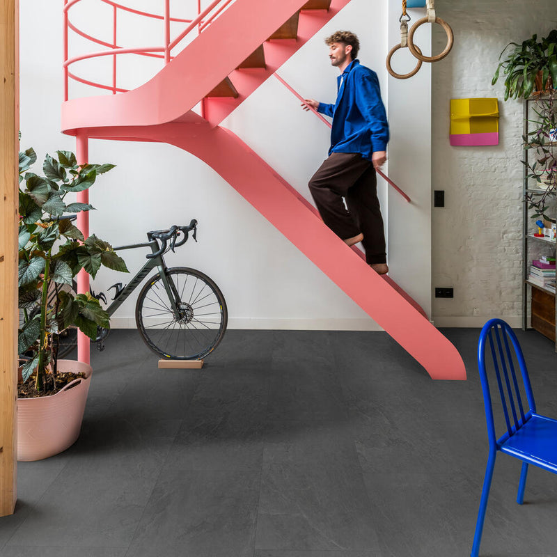 donkergrijze tegelvloer met matte structuur en lichte golvende lijnen, gecombineerd met een felroze spiraalvormige trap en diverse kleurrijke interieuraccenten zoals een blauwe stoel en een groene plant in een lichtroze pot