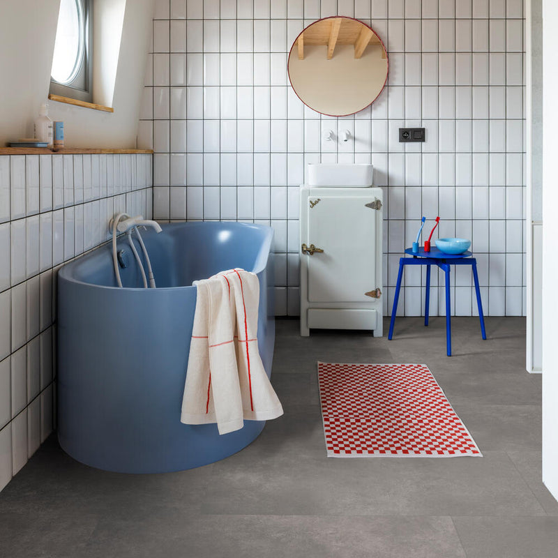 donkergrijze tegelvloer met grote, matte tegels met subtiel structuurpatroon in een badkamer met witte rechthoekige wandtegels, blauwe vrijstaande badkuip en blauw bijzettafeltje