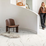 Witte tegelvloer met marmerpatroon in lichte beige en grijsbruine aders, brede tegels, moderne zithoek met donkerbruine houten stoel en lichtgrijze schapenvacht op de vloer.