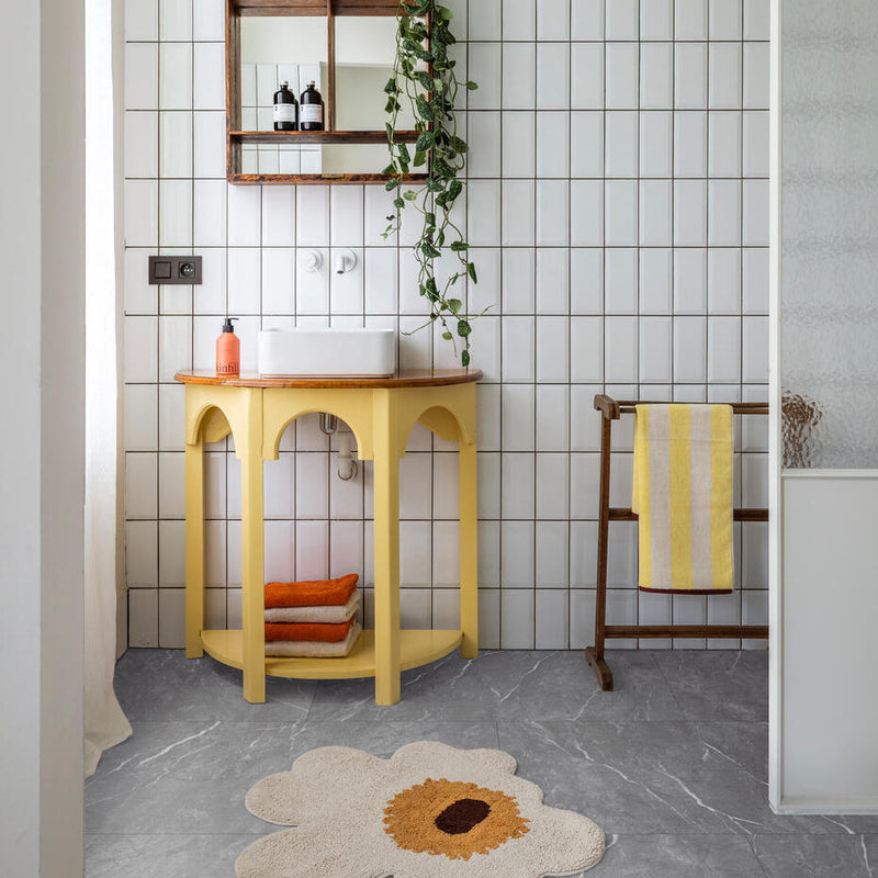 grijze tegelvloer met grote tegels in marmerstructuur, witte rechthoekige wandtegels in gridpatroon, gele houten badkamermeubel met afgeronde hoeken en open plank, beige en oranje opgevouwen handdoeken op onderste plank, witte wastafel met oranje zeeppompje, houten handdoekrek met gele en beige gestreepte handdoek, beige bloemvormige vloerkleed met bruin en okerkleurig midden